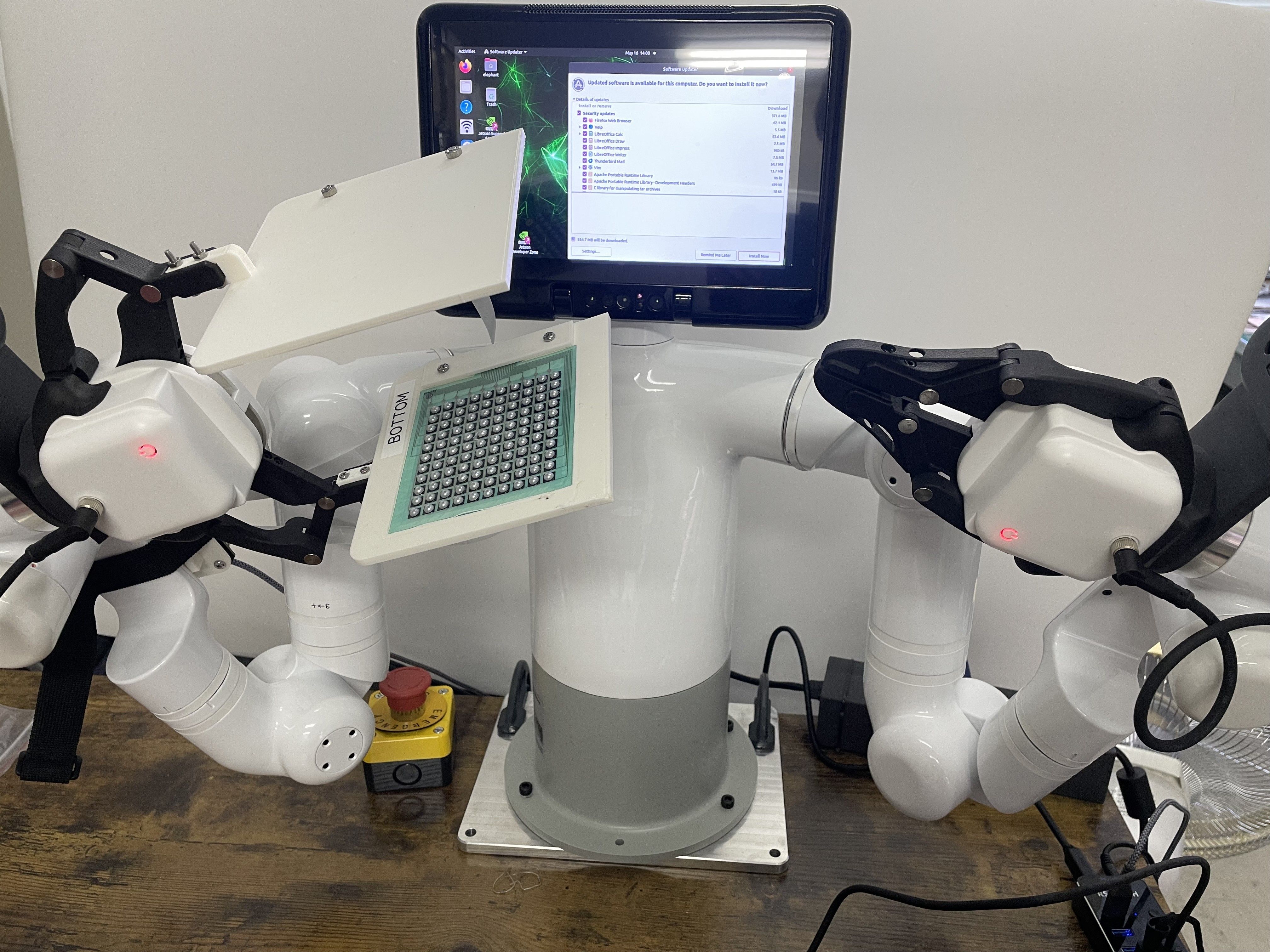 2本のアームを持つ白いロボットが、格子状にセンサが並んだ板を持っている様子。後方にモニターがある