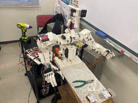 作業台の上で組み立て中の白いロボットハンドとアーム、周囲に配線と工具がある